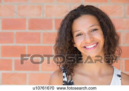 Stock Image - Portrait of happy girl in front of brick wall. Fotosearch