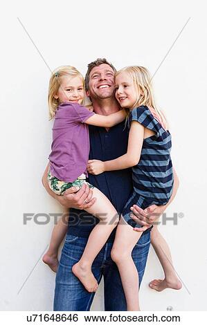 Stock Photograph - Portrait of mature man carrying two young daughters in front of white wall. Fotosearch