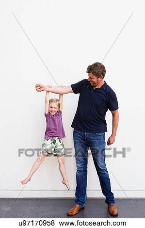 Stock Photo - Portrait of mature man with daughter hanging from his arm in front of white wall. Fotosearch
