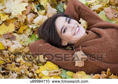 Stock Image - Portrait of mid adult woman lying amongst autumn leaves in park. Fotosearch