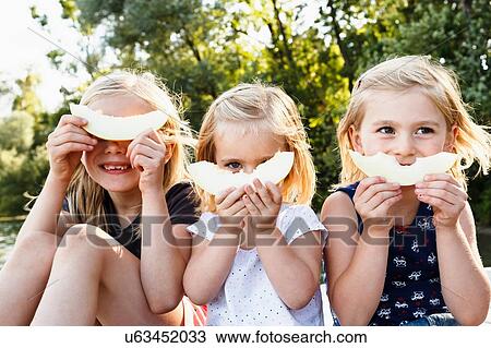 Stock Image - Portrait of three young sisters holding smiling melon in front of face in park. Fotosearch
