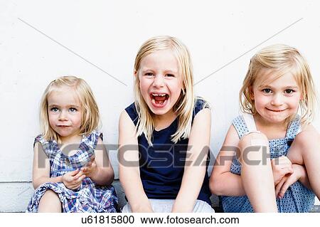 Stock Image - Portrait of three young sisters sitting in front of white wall. Fotosearch