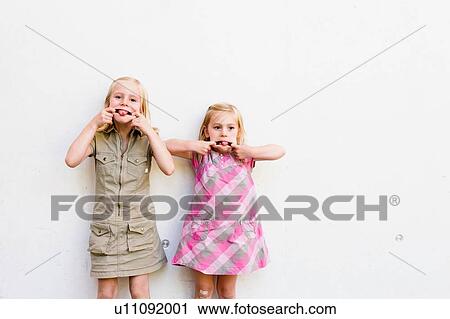 Portrait of two cute sisters pulling faces in front of white wall View Large Photo Image Stock Image - Portrait of two cute sisters pulling faces in front of white wall. Fotosearch