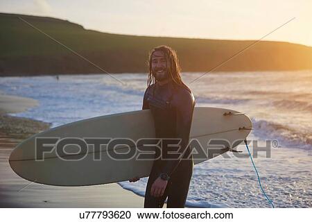 Stock Image - Portrait of young male surfer carrying surfboard on beach, Devon, England, UK. Fotosearch