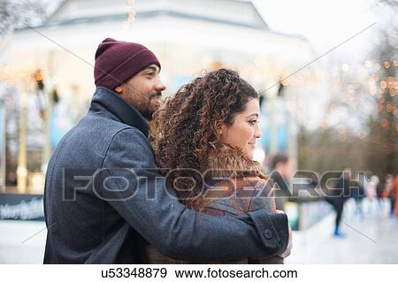 Stock Photo - Rear view mid adult man wearing knit hat with arm around young woman, looking away smiling. Fotosearch