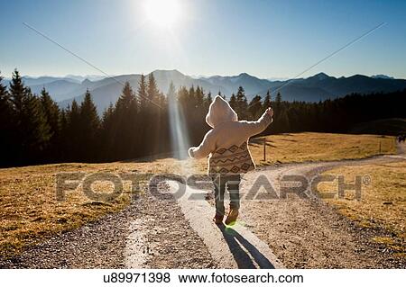 Stock Photo - Rear view of female toddler toddling on sunlit dirt track, Tegernsee, Bavaria, Germany. Fotosearch