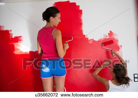 Stock Image - Rear view of girl and mother painting red wall with paint rollers. Fotosearch