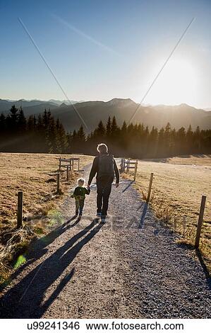 Stock Photograph - Rear view of man and toddler daughter walking on dirt track, Tegernsee, Bavaria, Germany. Fotosearch