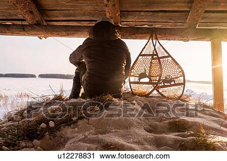 Stock Image - Rear view of man sitting on snowy haystack in barn next to snow shoes, Ural, Russia. Fotosearch