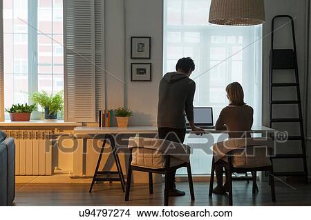 Rear view of mid adult couple using laptop at table in front of window View Large Photo Image Picture - Rear view of mid adult couple using laptop at table in front of window. Fotosearch