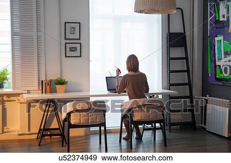 Rear view of mid adult woman using laptop at table in front of window View Large Photo Image Stock Photo - Rear view of mid adult woman using laptop at table in front of window. Fotosearch