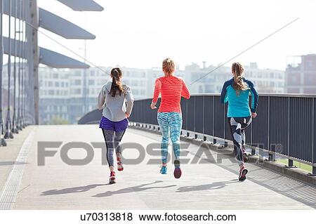 Stock Photo - Rear view of three female runners running across city footbridge. Fotosearch