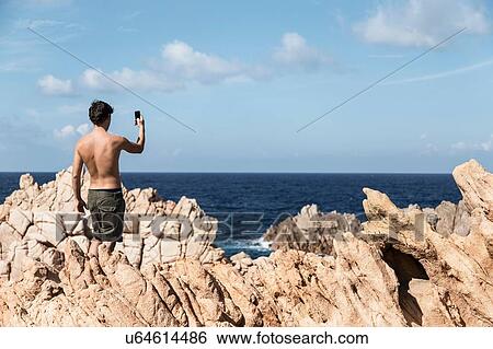 Stock Photograph - Rear view of young man on rock using smartphone to take photograph of horizon over ocean, Costa Paradiso, Sardinia, Italy. Fotosearch