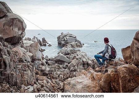 Rear view of young man sitting on rocks looking at view of ocean, Costa Smeralda, Sardinia, Italy View Large Photo Image Stock Image - Rear view of young man sitting on rocks looking at view of ocean, Costa Smeralda, Sardinia, Italy. Fotosearch