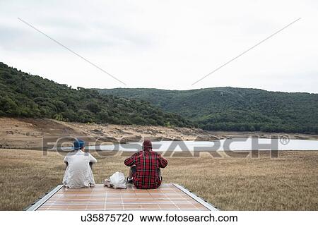 Stock Image - Rear view of young men sitting on wooden pier looking at mountain range, Nuoro, Sardinia, Italy. Fotosearch