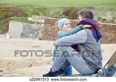 Stock Image - Romantic young couple sitting on car hood at beach. Fotosearch
