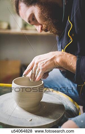 Stock Photo - Side view of bearded mid adult man looking down, making clay pot on pottery wheel. Fotosearch