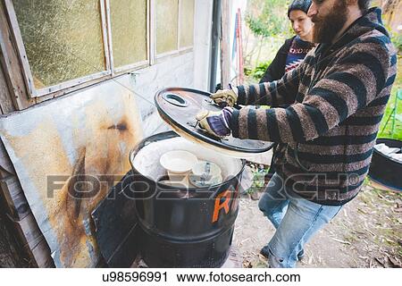 Stock Image - Side view of couple by shed lifting lid of barrel kiln. Fotosearch