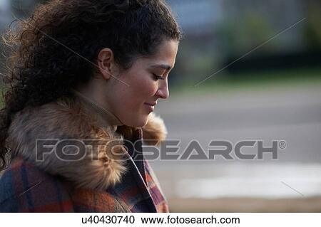Stock Image - Side view of curly haired woman wearing tartan fur trim coat looking down smiling. Fotosearch
