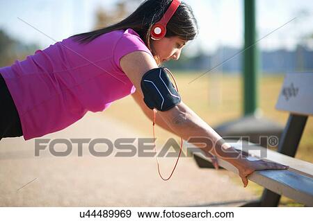 Stock Photo - Side view of mature woman wearing headphones doing push up using park bench. Fotosearch