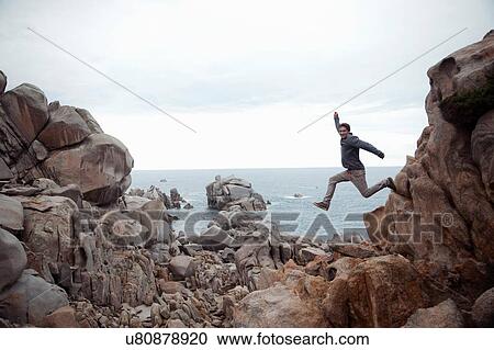 Side view of young man in mid air leaping from rocks, looking at camera, smiling, Costa Smeralda, Sardinia, Italy View Large Photo Image Stock Image - Side view of young man in mid air leaping from rocks, looking at camera, smiling, Costa Smeralda, Sardinia, Italy. Fotosearch