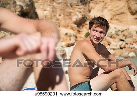 Side view of young man sitting on beach with friend smiling, Golfo di Orosei, Sardinia, Italy View Large Photo Image Stock Image - Side view of young man sitting on beach with friend smiling, Golfo di Orosei, Sardinia, Italy. Fotosearch