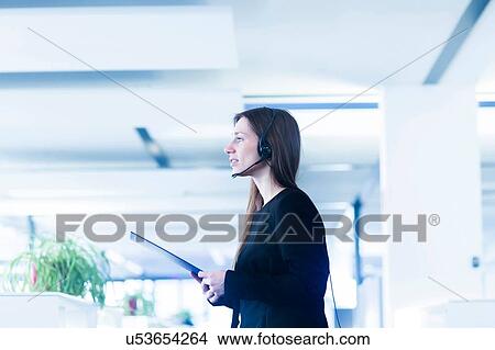 Picture - Side view of young woman in office wearing telephone headset, holding digital tablet looking away. Fotosearch