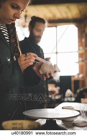 Stock Image - Side view of young woman in workshop brushing clay pot. Fotosearch