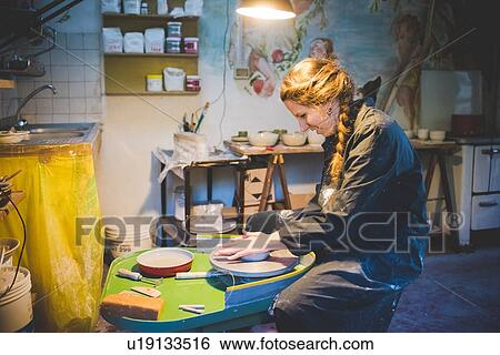 Side view of young woman sitting in workshop using potters wheel, looking down smiling View Large Photo Image Stock Photograph - Side view of young woman sitting in workshop using potters wheel, looking down smiling. Fotosearch