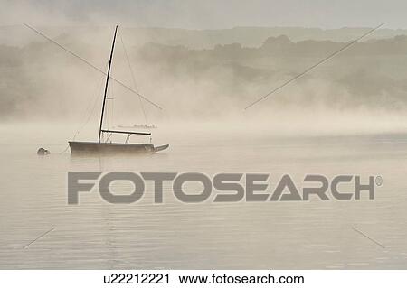Stock Image - Silhouetted yacht on misty Wimbleball lake at dawn, Exmoor, Somerset, England. Fotosearch