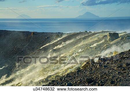 Stock Image - Sulphur and fumaroles smoke on Gran Cratere; The Islands of Alicudi and Filicudi in the background, Vulcano Island, Aeolian Islands, Sicily, Italy. Fotosearch
