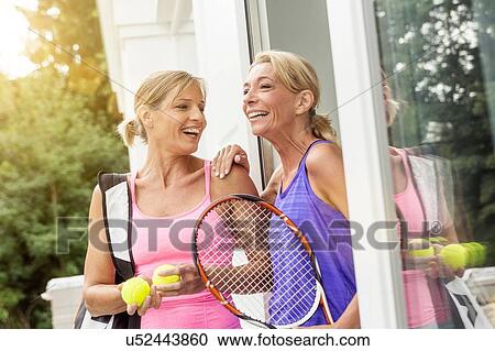 Stock Image - Two mature women preparing to play tennis at patio door. Fotosearch