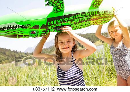 Stock Image - Two young girls walking through field, carrying inflatable crocodile. Fotosearch