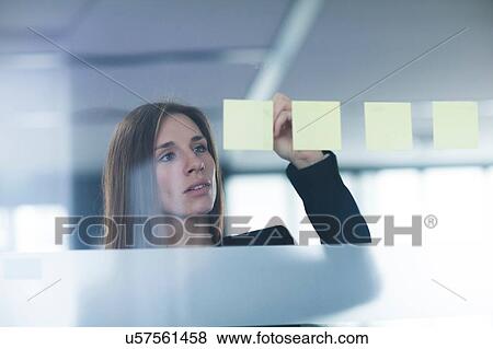 Stock Photo - View through window of young woman writing on post it note stuck to glass. Fotosearch