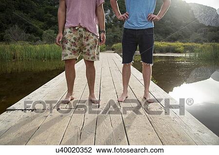 Stock Image - Waist down front view of young men on wooden pier, Cala Luna, Sardinia, Italy. Fotosearch