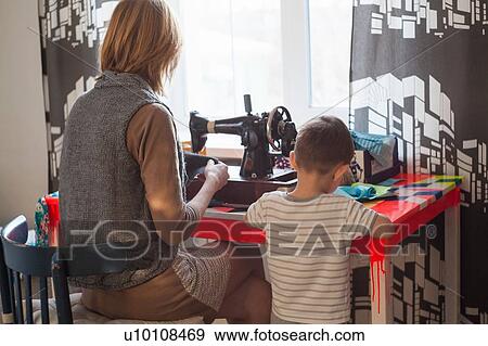 Stock Photo - Woman and toddler son using vintage sewing machine at table in front of window. Fotosearch