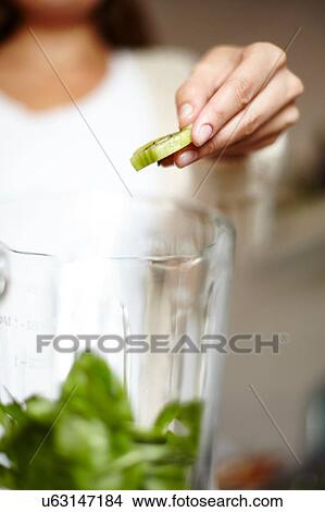 Picture - Woman holding sliced green kiwi over blender. Fotosearch