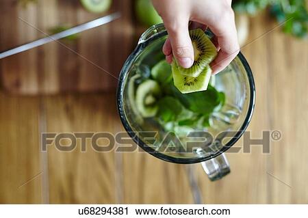 Stock Image - Woman holding sliced green kiwi over blender. Fotosearch
