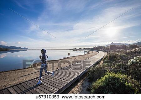 Stock Photo - Young female runner running on beach boardwalk, Villasimius, Sardinia, Italy. Fotosearch