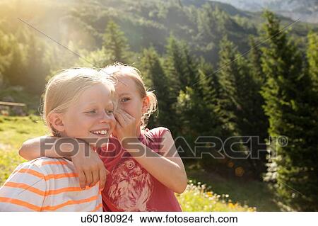 Picture - Young girl whispering in friends ear, outdoors. Fotosearch