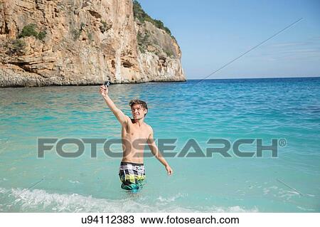 Stock Image - Young man in ocean arm raised using camera smiling, Golfo di Orosei, Sardinia, Italy. Fotosearch