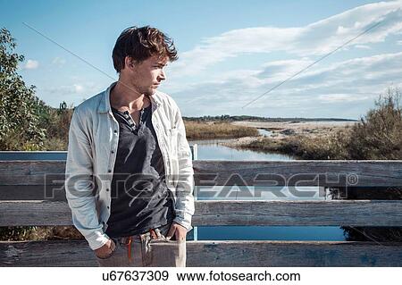 Young man leaning against wooden fence on bridge over river, hands in pockets looking away, Costa Smeralda, Sardinia, Italy View Large Photo Image Stock Photo - Young man leaning against wooden fence on bridge over river, hands in pockets looking away, Costa Smeralda, Sardinia, Italy. Fotosearch