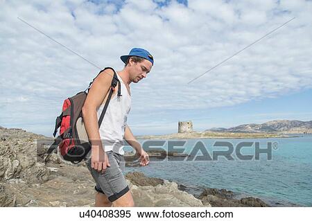 Stock Photography - Young man on rocks by ocean carrying backpack looking down, Stintino, Sardinia, Italy. Fotosearch
