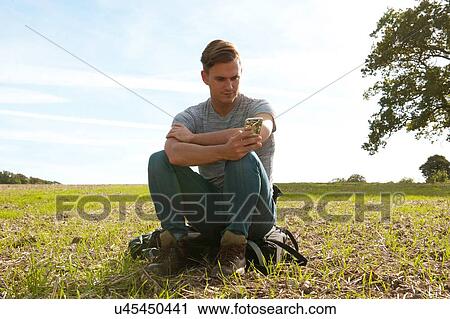 Young man sitting on backpack reading smartphone texts in field View Large Photo Image Stock Image - Young man sitting on backpack reading smartphone texts in field. Fotosearch
