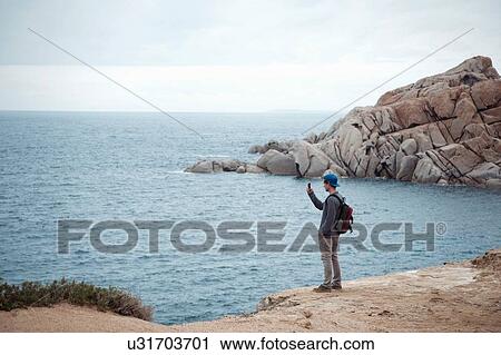 Stock Image - Young man standing on cliff using smartphone to photograph ocean, Costa Smeralda, Sardinia, Italy. Fotosearch
