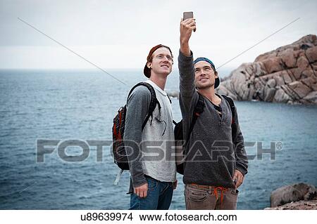 Young men in front of ocean using smartphone to take selfie smiling, Costa Smeralda, Sardinia, Italy View Large Photo Image Picture - Young men in front of ocean using smartphone to take selfie smiling, Costa Smeralda, Sardinia, Italy. Fotosearch