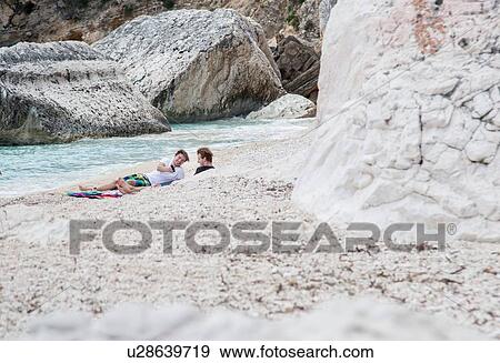 Stock Photo - Young men relaxing on rocky beach using smartphone, Golfo di Orosei, Sardinia, Italy. Fotosearch