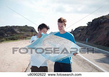 Stock Image - Young men standing on road looking at map smiling, Costa Smeralda, Sardinia, Italy. Fotosearch