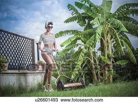 Stock Image - Young stylish woman pushing lawnmower in garden. Fotosearch