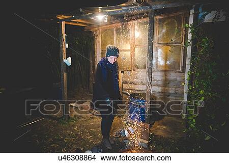 Young woman by shed waving tongs over fire causing sparks, looking down smiling View Large Photo Image Picture - Young woman by shed waving tongs over fire causing sparks, looking down smiling. Fotosearch